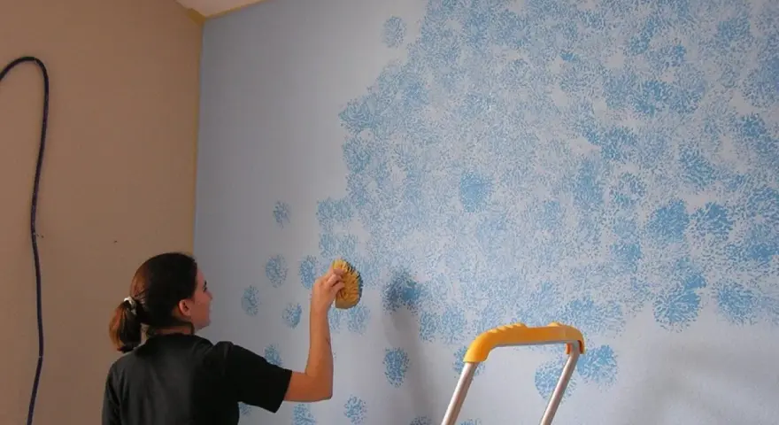 Mujer aplica pintura que disimula imperfecciones en pared con esponja, creando un patrón de nubes azules.