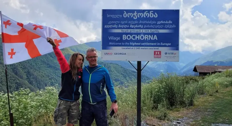 Para, z gruzińską flagą, na tle gór i znaku "Wioska Bochorna", najwyżej położona osada w Europie. Podróżując, warto zapytać: czy w Gruzji jest bezpiecznie?
