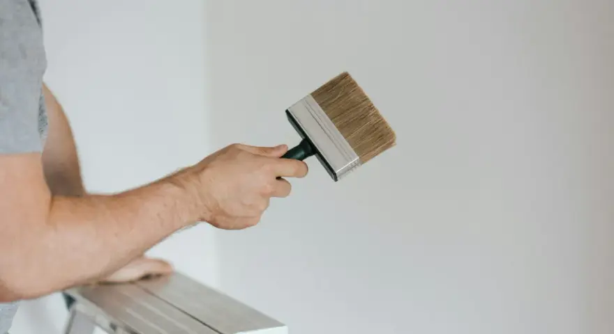 Person på en stege håller en bred pensel redo att måla huset vitt.