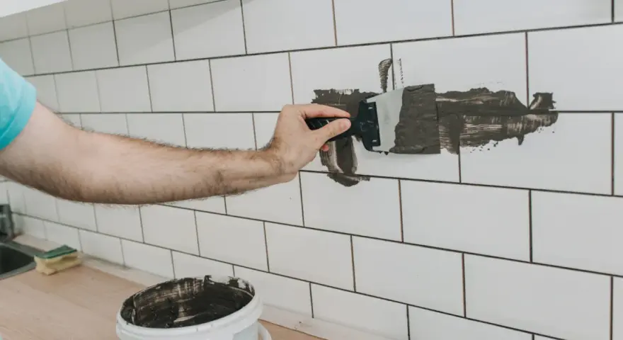 Ręka nakłada ciemną zaprawę na białe płytki, pokazując, jak zmienić kolor fugi na ciemniejszy. Wiadro z zaprawą stoi obok.