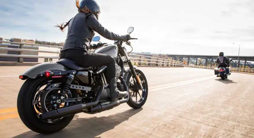 Kobieta w skórzanej kurtce i kasku jedzie na stylowym motocyklu dla kobiety. Wiatr rozwiewa jej włosy, a słońce odbija się od chromowanych elementów.
