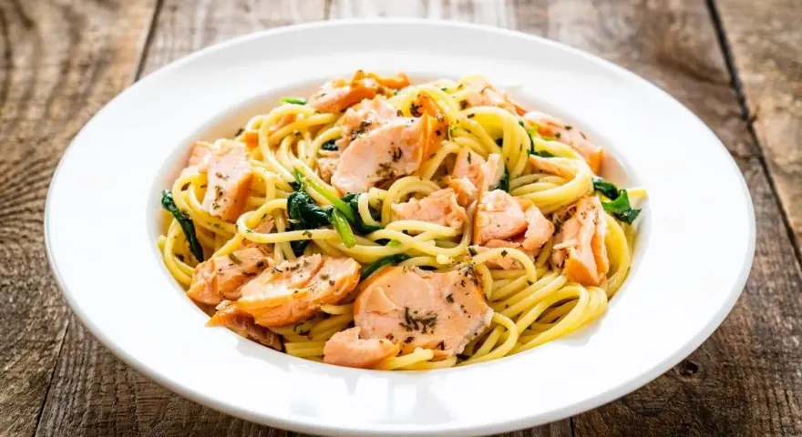 Makaron spaghetti z łososiem i szpinakiem na białym talerzu, na drewnianym tle.