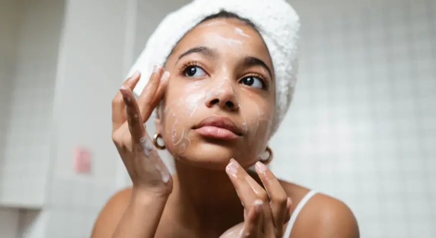 Una joven con toalla en la cabeza se aplica espuma en el rostro, parte esencial de su rutina de skincare.