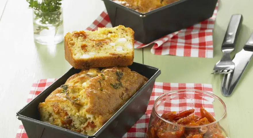 Cake tomates séchées feta, un délice salé aux herbes fraîches. Une tranche est posée sur un cake encore dans son moule.
