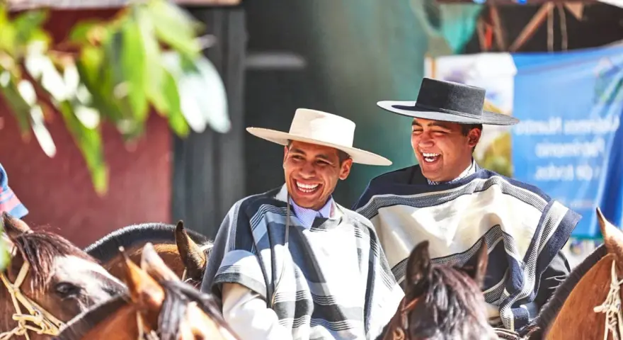Deux huasos rient joyeusement à cheval, vêtus de ponchos traditionnels et de chapeaux.