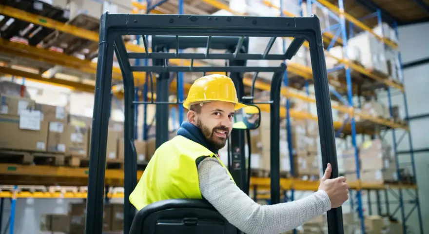 Uśmiechnięty operator wózka widłowego w żółtym kasku i kamizelce odblaskowej, z kciukiem w górze. Zastanawiasz się, ile zarabia operator wózka widłowego?