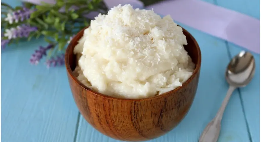 Una ciotola di legno piena di cremosa crema al cocco, guarnita con cocco grattugiato. Accanto, un cucchiaino d'argento e fiori di lavanda.