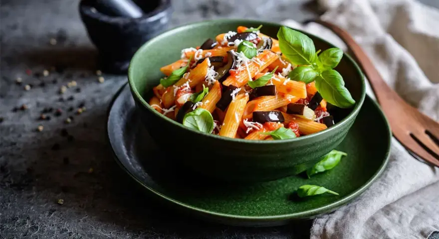 Un plato de pasta alla norma, con penne, berenjena, salsa de tomate y albahaca fresca, espolvoreado con queso rallado.
