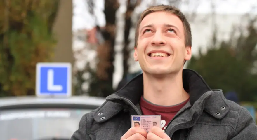 Szczęśliwy młody mężczyzna trzyma prawo jazdy kategorii B, z niebieskim znakiem "L" na dachu samochodu w tle.