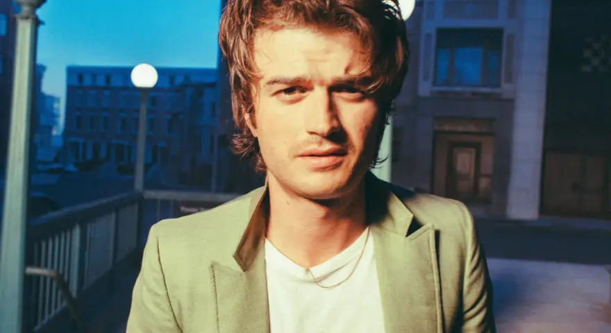 Joe Keery w jasnej marynarce, z naszyjnikiem, na tle nocnego miasta.