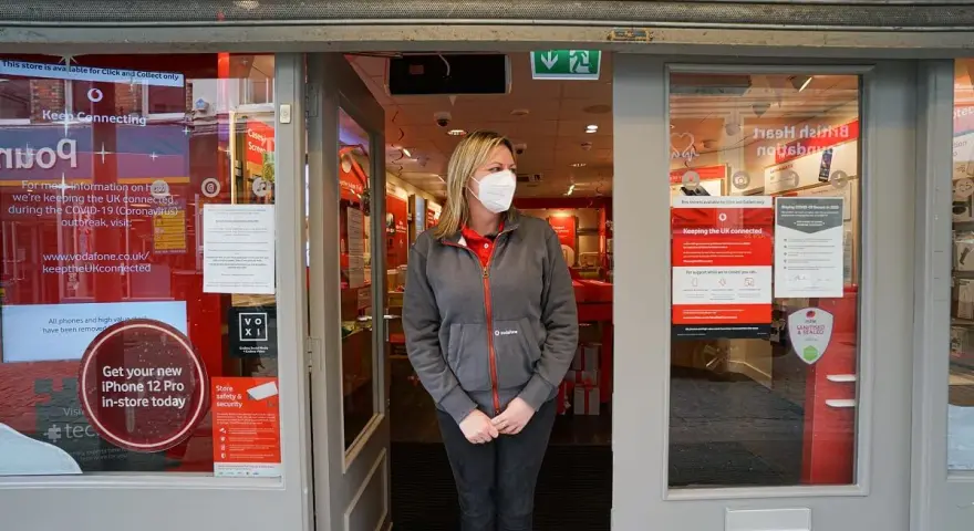 Kobieta w maseczce stoi przed zamkniętym sklepem Vodafone. Informacja na drzwiach głosi, że sklep jest otwarty tylko na odbiór zamówień online, nawet w bank holiday.