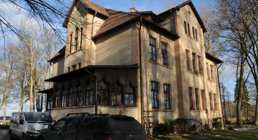 Dawny pałac Poraj, z charakterystyczną werandą i drzewami w tle, otoczony zimowym krajobrazem.