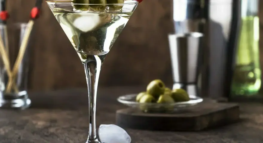 Elegancki martini drink z trzema oliwkami na patyczku, lodem i akcesoriami barmańskimi w tle.