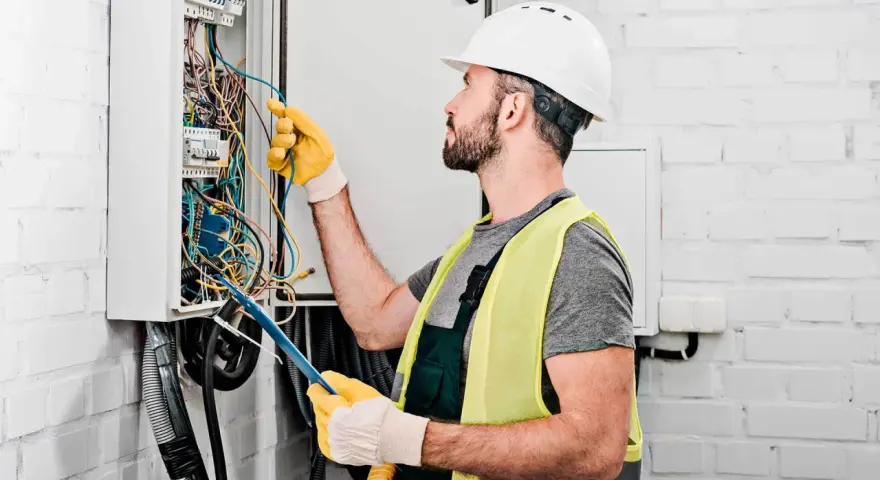 Elektryk w kasku i kamizelce odblaskowej pracuje przy skrzynce z przewodami. Sprawdź nasze usługi elektryczne cennik.
