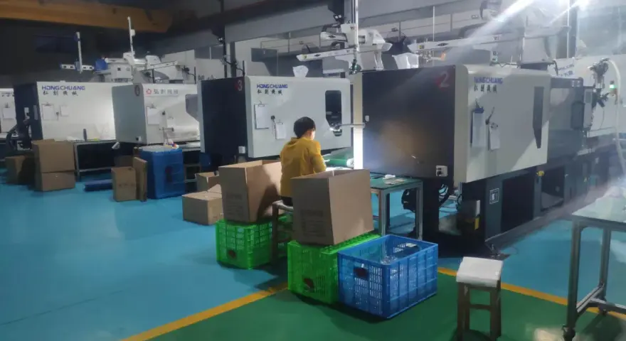 A person in a yellow jacket works at a station with boxes and crates, surrounded by injection molding machines, indicating low volume production.
