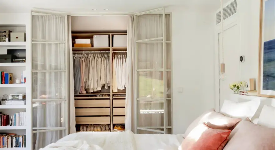 Dormitorio con vestidor integrado. Ropa organizada en armarios abiertos y estanterías. Cama con cojines rosas y manta blanca.