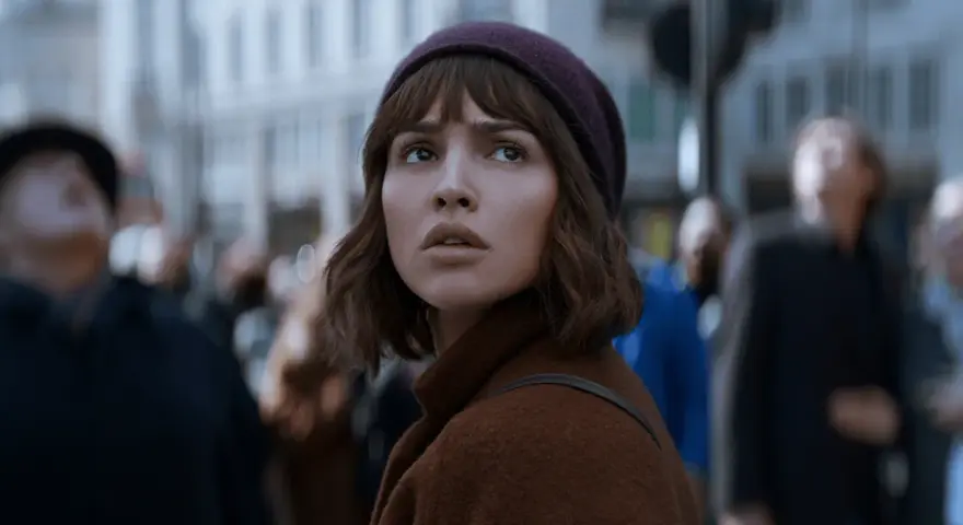 Młoda kobieta w brązowym płaszczu i bordowej czapce patrzy w górę, otoczona tłumem. Czy to zapowiedź netflix nowości dzisiaj?