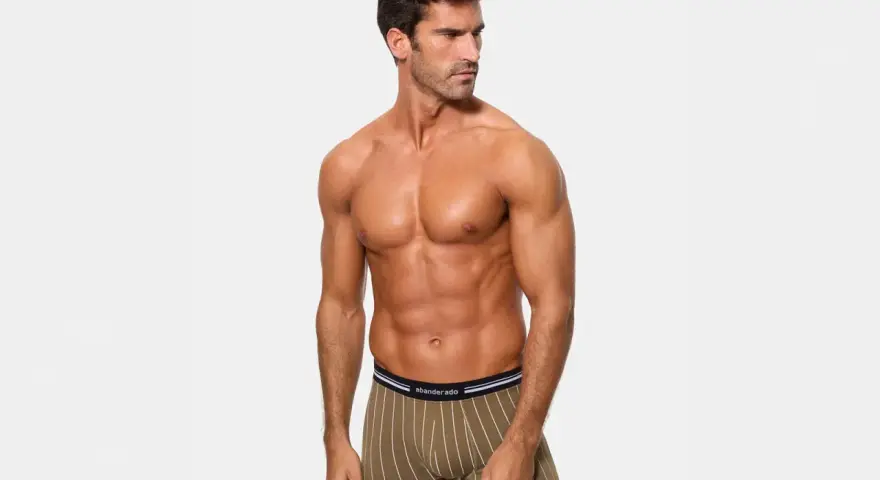 Hombre musculoso posando con calzoncillos de poliamida abriga, con rayas verticales blancas sobre fondo verde oliva.