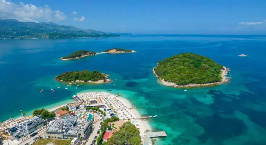 Rajska plaża z zielonymi wysepkami w **jeziorze w Albanii**. Woda jest turkusowa, a na piasku widać leżaki i budynki.