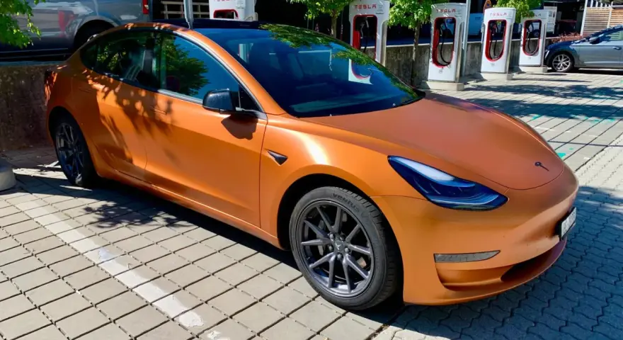 Pomarańczowa Tesla na tle ładowarek Supercharger.