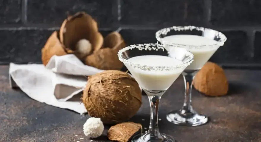 Dwa kieliszki drinka z malibu z mlekiem, ozdobione wiórkami kokosowymi, obok świeżych kokosów.