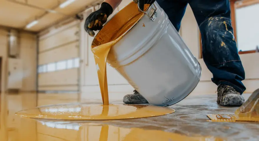 Hombre con guantes vierte pintura para suelo de cemento de color amarillo brillante de un cubo.