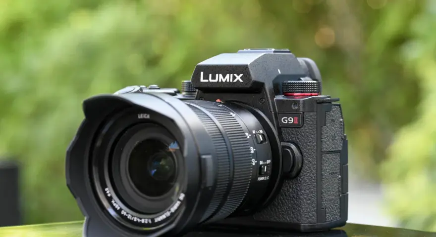 Un appareil photo noir Lumix G9 II avec objectif Leica est posé sur une surface réfléchissante, avec un arrière-plan flou de verdure.