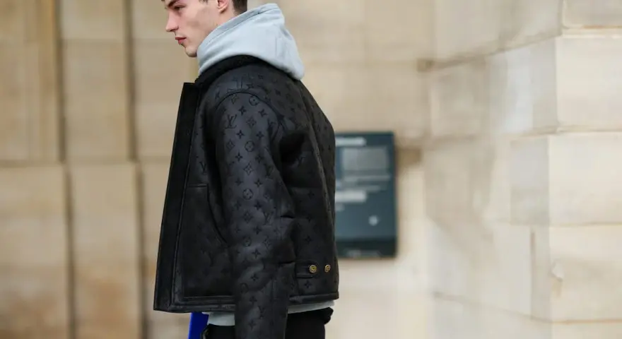 Uomo con un elegante outfit con giubbotto di pelle nero con monogrammi, felpa grigia e pantaloni scuri.