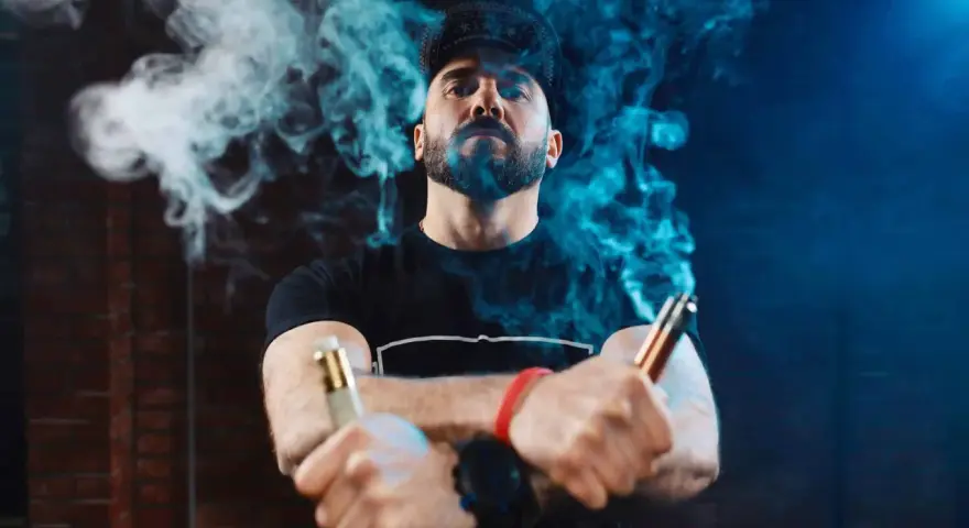 Hombre con barba y gorra, rodeado de vapor azul, sostiene dos vapers, uno dorado y otro cobre. La diferencia entre 0.8 y 1.2 ohm se siente en la calada.