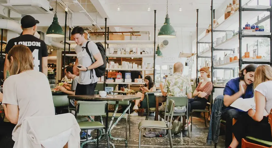 Gente en un café con mesas verdes y estanterías llenas de objetos. El **ruido restaurante** es palpable con las conversaciones y el movimiento.