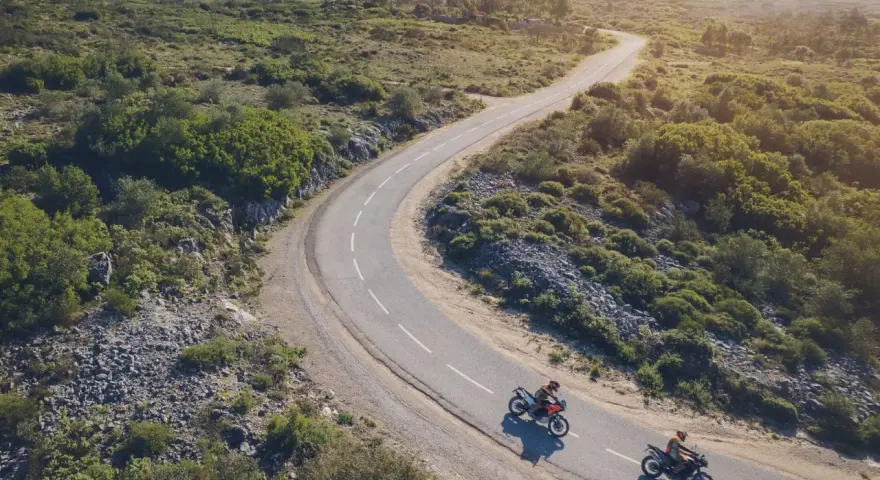 Due moto KTM sfrecciano su una strada tortuosa tra colline verdi. Un'avventura che celebra il KTM Connect.