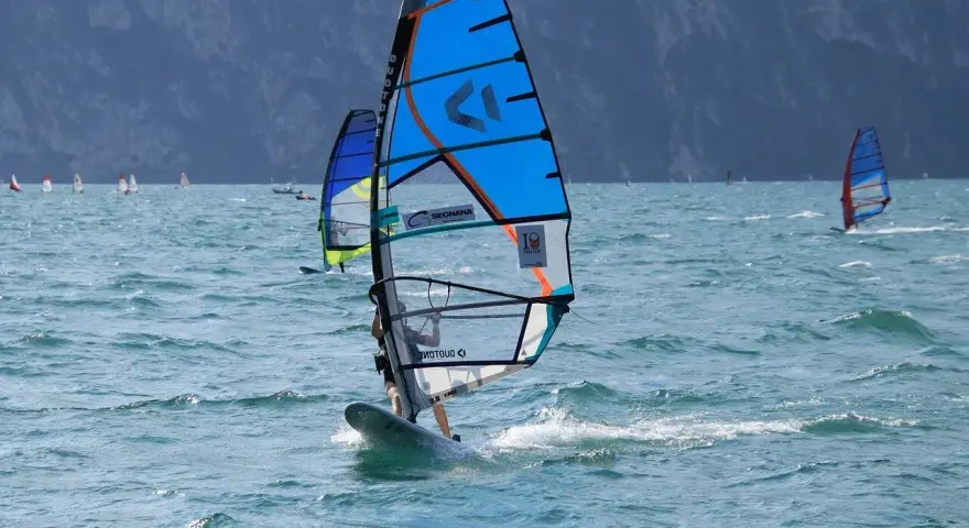 Osoba na desce windsurfingowej z niebieskim żaglem na wzburzonym morzu.