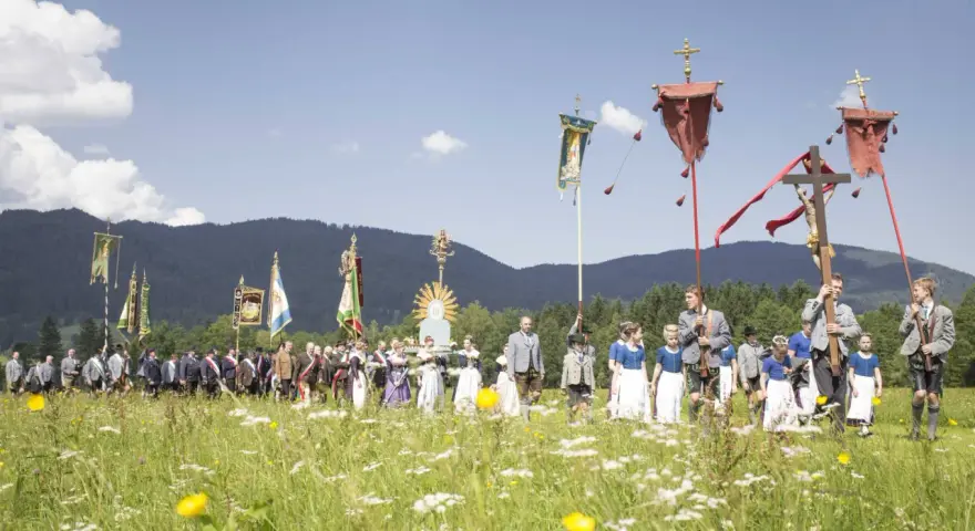 Menschen in Tracht und Uniformen nehmen an einer Prozession teil, die den Fronleichnam feiert. Fahnen und ein Kreuz werden getragen.