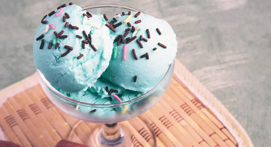 Ein Glas mit blauem Smurf-Eis, bestreut mit bunten Streuseln, steht auf einem Holzuntersetzer.