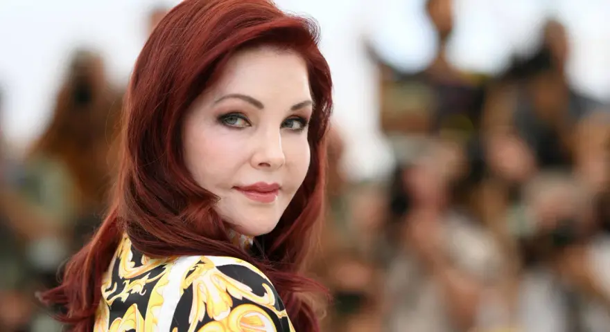Priscilla Presley, mit ihrem ikonischen roten Haar und einem Hauch von 70er-Jahre-Glamour, blickt nachdenklich in die Ferne.