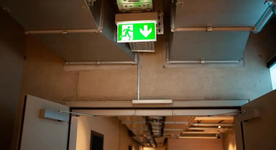 Zielone światła awaryjne wskazują drogę ewakuacji w korytarzu z widocznymi instalacjami wentylacyjnymi i betonowymi ścianami.
