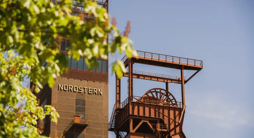 Industriearchitektur im Nordsternpark Gelsenkirchen: Ein markanter Förderturm und das Gebäude mit der Aufschrift "NORDSTERN" vor blauem Himmel.