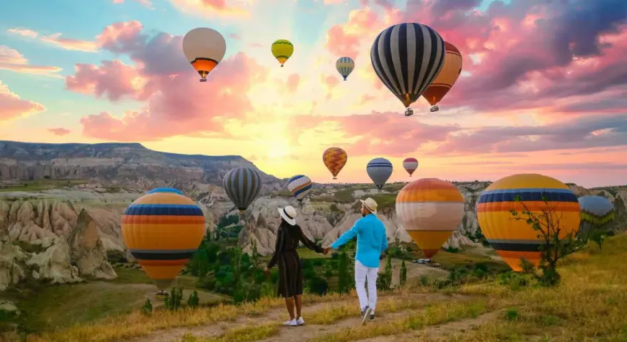 A couple holds hands, gazing at a sky filled with hot air balloons over the unique landscape of western Turkey at sunrise.