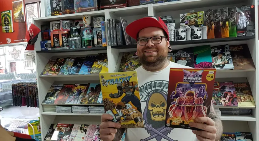 Mann mit Brille und roter Kappe hält zwei Comics in einem Comicladen.