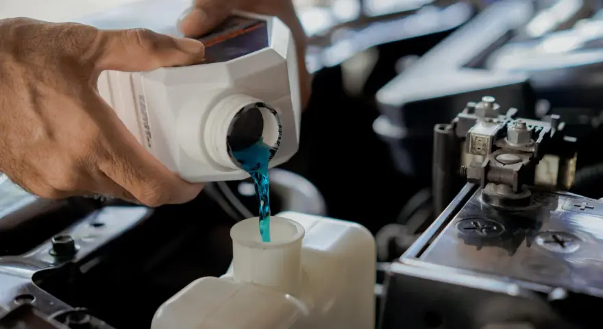 Hands pour blue coolant into a car's reservoir. This maintenance task is essential for preventing overheating, which can lead to costly car radiator replacement.