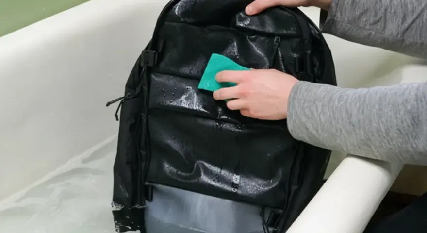 Se puede lavar una mochila en la lavadora. Alguien limpia una mochila negra en una bañera con agua y jabón.