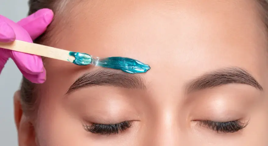 A pink-gloved hand applies turquoise wax to an eyebrow. This close-up shows the contrast between hot wax vs cold wax application for brow shaping.