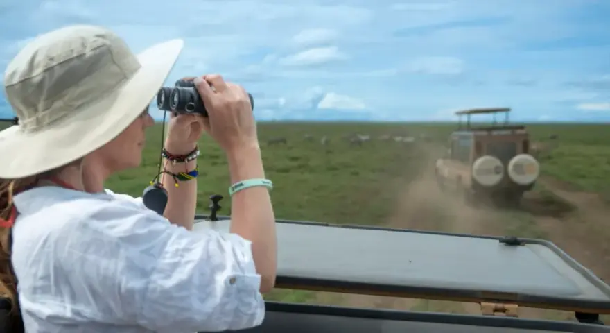 Mujer con sombrero y binoculares observa cebras en la sabana, disfrutando de su viaje a Tanzania.
