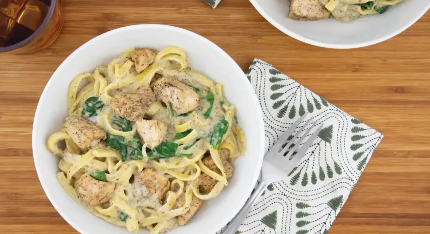 Delicioso plato de pasta alfredo con trozos de pollo y espinacas, servido en un tazón blanco sobre una tabla de madera.