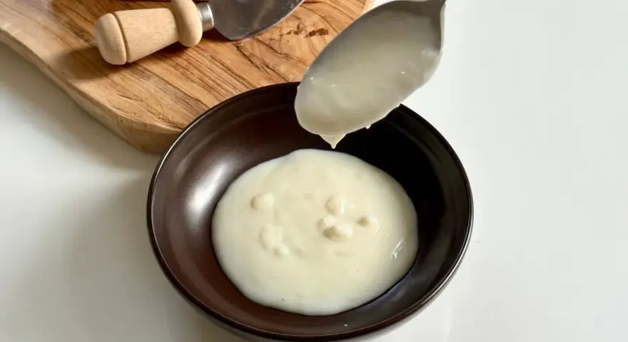 Una cucchiaiata di cremosa fonduta di parmigiano cade in una ciotola. Sul tagliere, un pezzo di Parmigiano Reggiano e un coltellino.