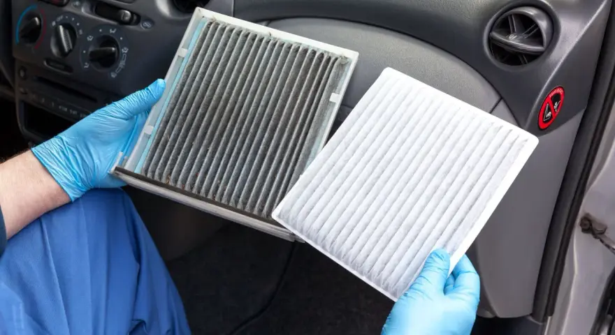 A dirty cabin air filter can make your car smell. A gloved hand holds a very dirty filter next to a clean, white one, showing the stark contrast.