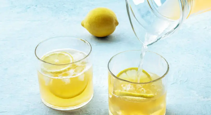 Erfrischendes zitronenwasser am morgen wird in ein Glas mit Eis und Zitronenscheiben gegossen. Eine ganze Zitrone liegt daneben.