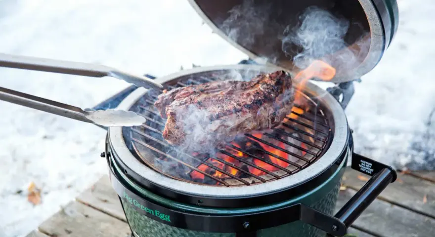 Un succulento bistec a la fiorentina cuoce sulla griglia di un Big Green Egg, circondato da fumo e fiamme.