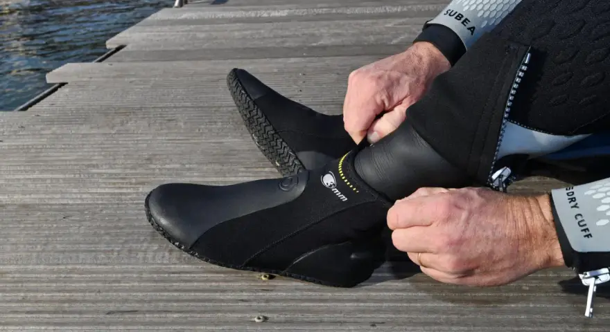 Mężczyzna zakłada czarne buty neoprenowe na drewnianym pomoście przy wodzie.
