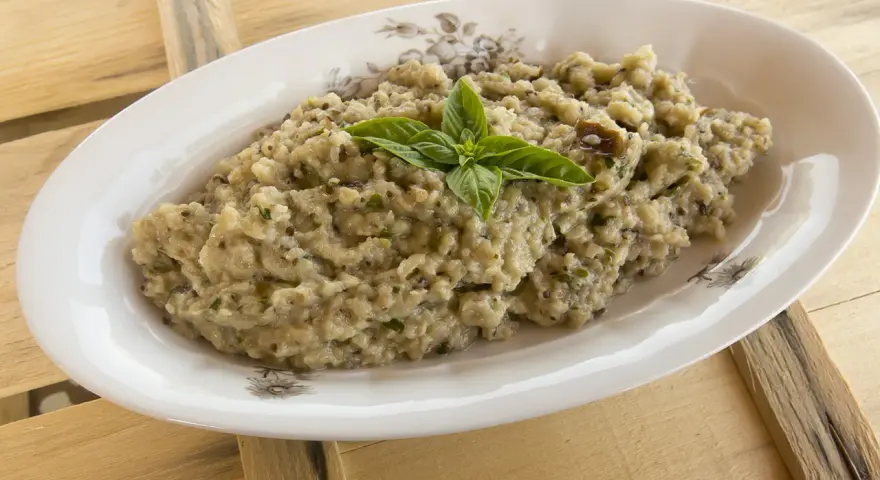 Hummus di melanzane cremoso, guarnito con basilico fresco, servito in un piatto ovale su un tavolo di legno grezzo.